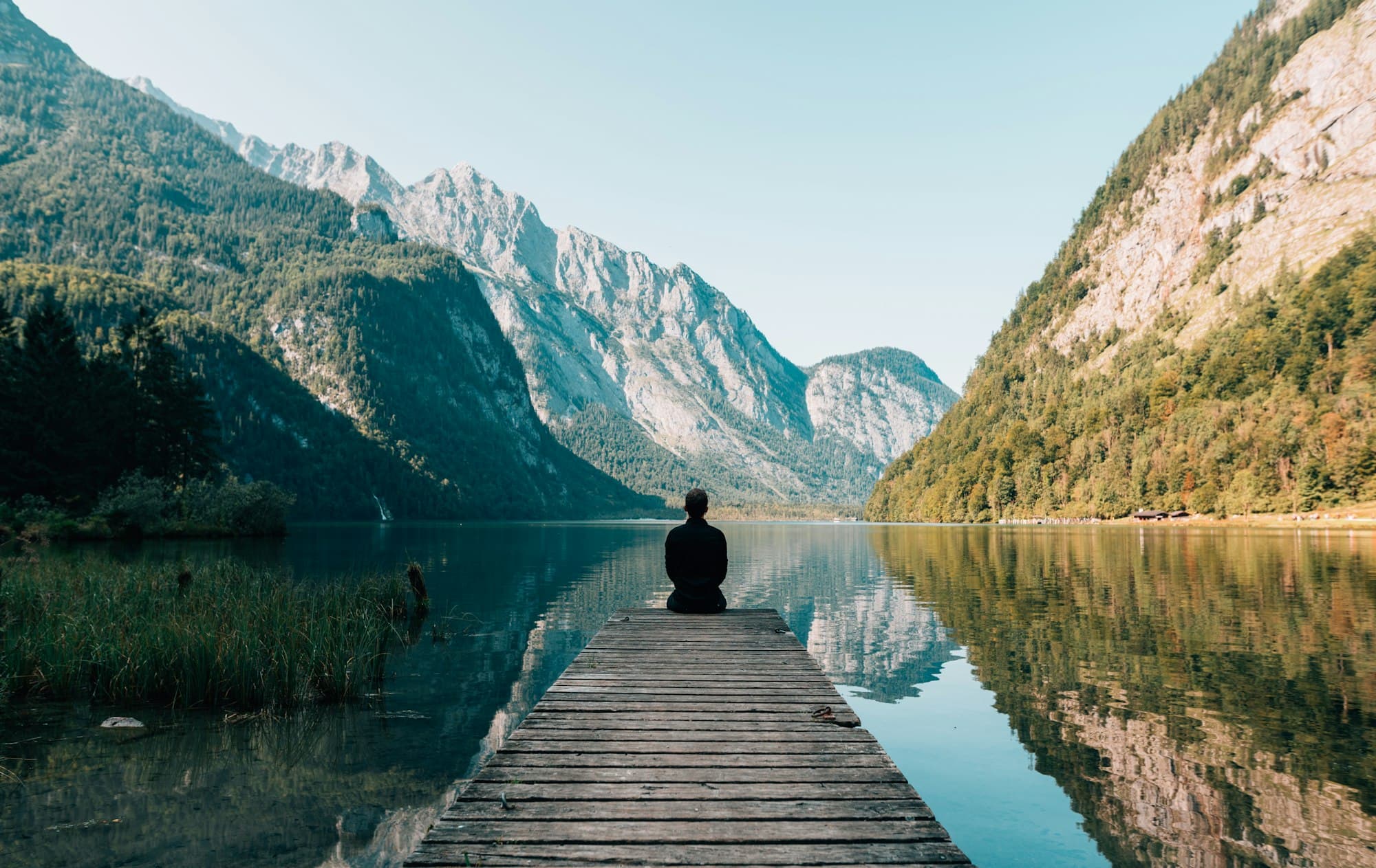 A person sitting at the end of a wooden dock on a still mountain lake